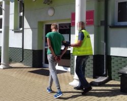 Policjant w kamizelce odblaskowej z napisem policja prowadzi zatrzymanego. W tle budynek Prokuratury Rejonowej w Biłgoraju.