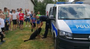 policjant z psem służbowym stoi przy radiowozie, obok stoją dzieci z opiekunami