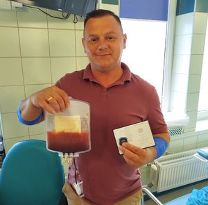 policjant po zabiegu oddania komórek macierzystych stoi w szpitalnej sali trzymając  w jednej dłoni legitymację dawcy przeszczepu a w drugiej worek z krwi