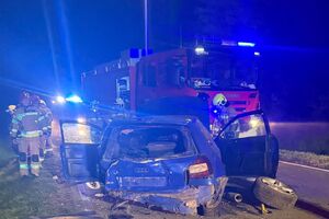 rozbity samochód na drodze, w tle wóz strażacki i strażacy